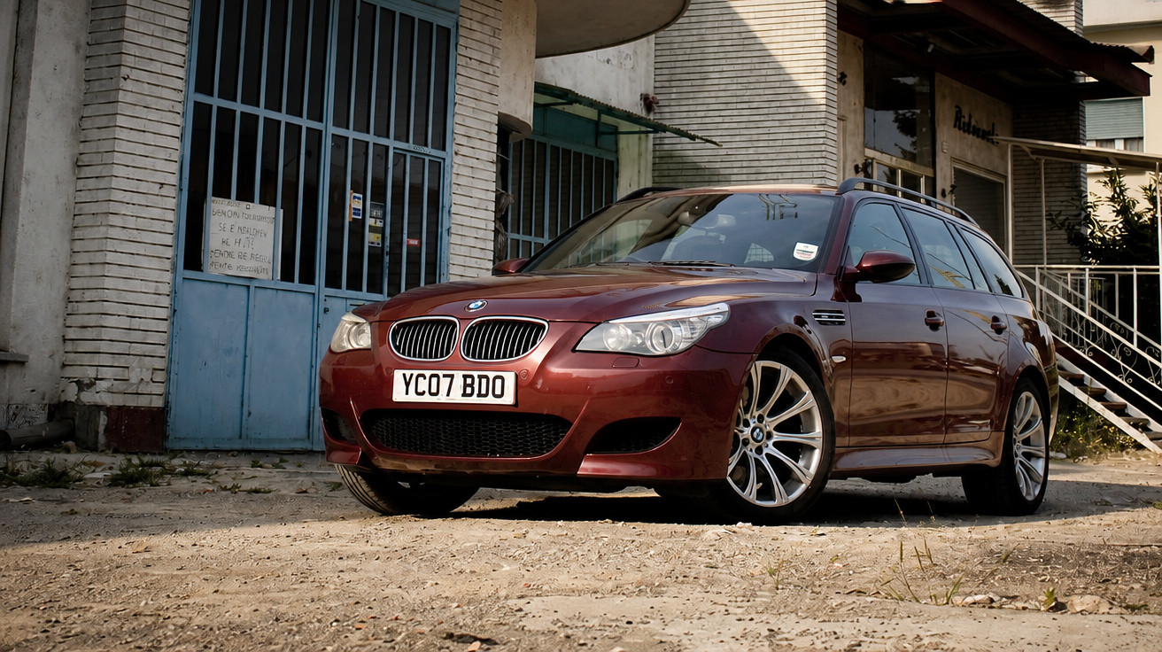 Automóvel BMW Touring vermelho estacionado em chão de terra perto de edifício antigo.